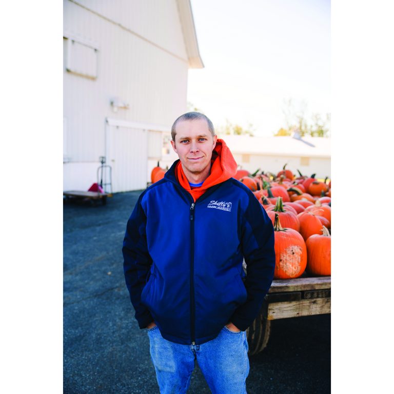 Scott Skelly smiles in front of a wagon filled with pumpkins.