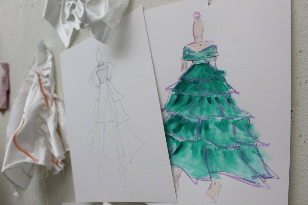A design sketch of a dress hangs on the wall of a studio next to fabric samples.